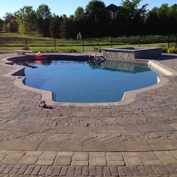 A wide view of a luxury inground swimming pool with a unique, irregular shape, surrounded by textured stone pavers. The pool area includes a raised stone hot tub, a diving board, and metal steps, set against a background of a green lawn, black wrought-iron fence, and mature trees.