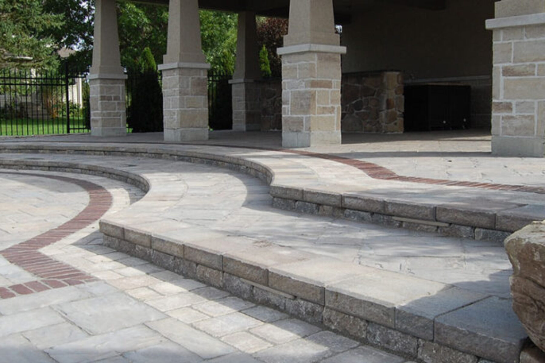 A multi-level outdoor stone patio featuring grey pavers with a curved red brick inlay. The space includes a covered pavilion supported by stone columns.