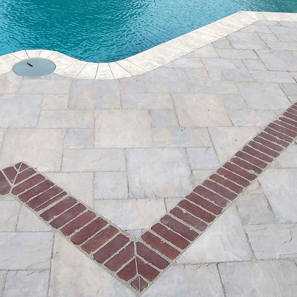 A close-up, overhead view of a swimming pool deck featuring light grey textured stone pavers. A decorative border of dark red bricks creates a sharp V-shaped inlay, leading toward the edge of a turquoise pool with light-colored coping.