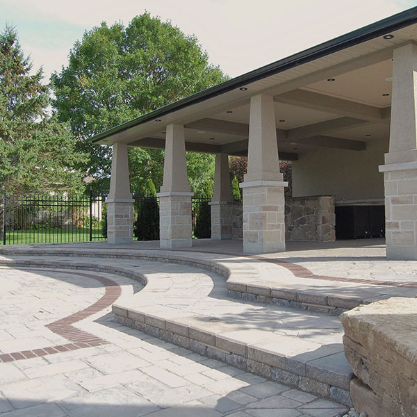 A multi-level outdoor stone patio featuring grey pavers with a curved red brick inlay. The space includes a covered pavilion supported by stone columns.