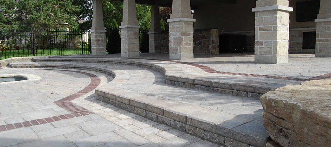 A multi-level outdoor stone patio featuring grey pavers with a curved red brick inlay. The space includes a covered pavilion supported by stone columns.